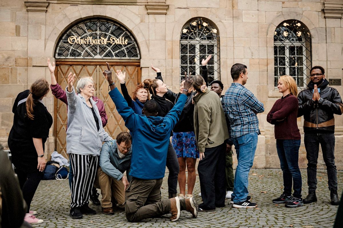 Esslinger Suppkultur bei "Stadt im Fluss" © Sabine Watzko Eine Gruppe von Menschen unterschiedlichen Alters steht vor einem historischen Gebäude und führt gemeinsam eine Art Bewegungs- oder Theaterübung aus, wobei mehrere Personen die Arme nach oben strecken, während andere zuschauen oder sich abwenden.