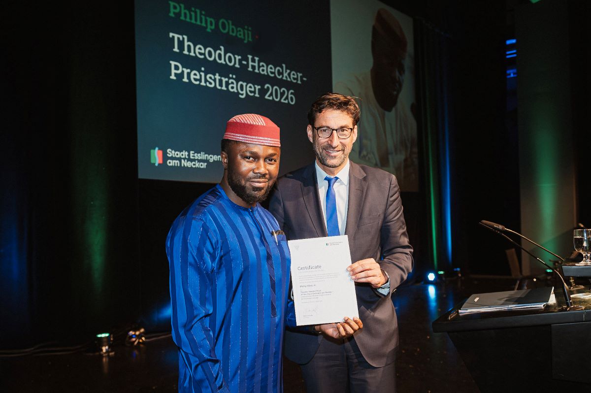 THP2026 Obaji-Urkunde ©Sabine Watzko Zwei Männer stehen auf einer Bühne und halten gemeinsam eine Urkunde. Im Hintergrund ist eine große Leinwand mit Text zur Preisverleihung „Theodor-Haecker-Preisträger 2026“ zu sehen.