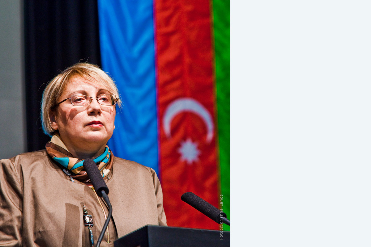 Dr. Leyla Yunus © Krishna Lahoti Frau mit kurzen, blonden Haaren und Brille in brauner Jacke mit Halstuch spricht am Rednerpult in ein Mikrofon. Im Hintergrund eine Flagge mit blauem, rotem und grünem Streifen. Auf dem roten sind ein Halbmond und ein Stern zu sehen.