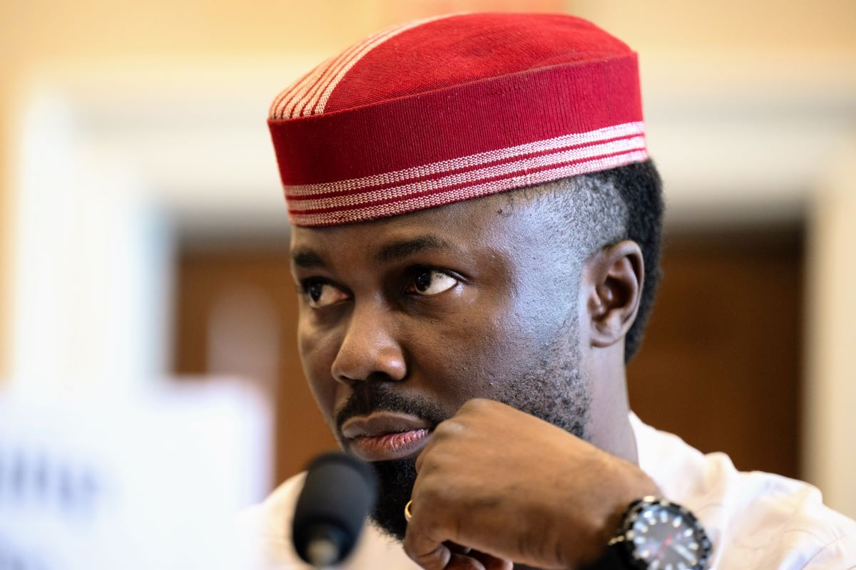 Philip Obaji jr., mit freundlicher Genehmigung der U.S. Helsinki Commission Portrait eines Mannes in weißer Kleidung und rot-weißem traditionellem Kopfschmuck, mit gepflegtem Bart, die Hand am Kinn ruhend, vor einem Mikrofon in einem Innenraum, aufmerksam und nachdenklich.