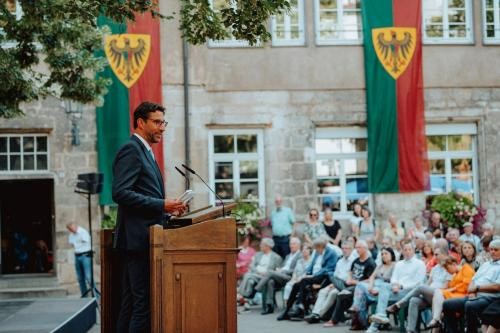 Mann am Redepult spricht zu sitzendem Publikum im Freien. Am Gebäude im Hintergrund hängen grün-rote Fahnen mit gelbem Wappen, auf denen ein Adler abgebildet ist.