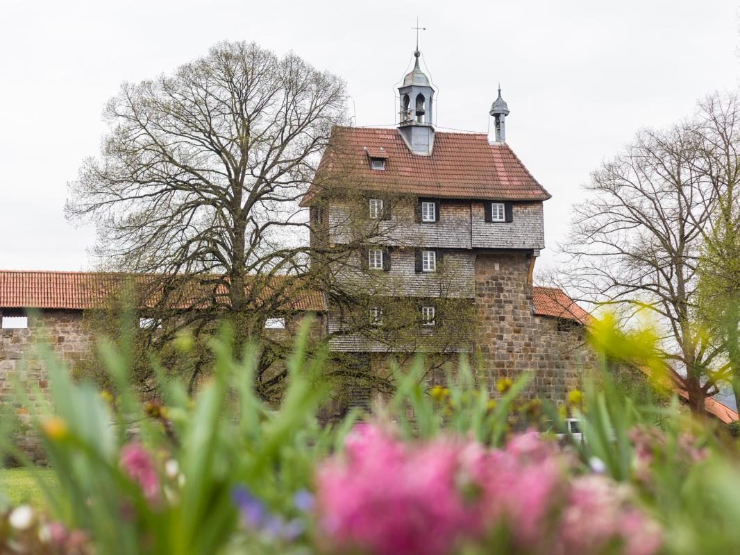 Schindelbedecktes Gebäude mit Glockentürmchen auf einer Mauer. Im Vordergrund Blumen.