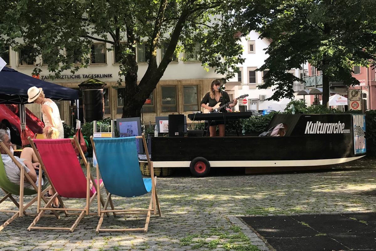 Auf dem schwarzen Holzboot "Kulturarche" steht eine Sängerin am Mikrofon. Davor sitzen Personen in Liegestühlen.