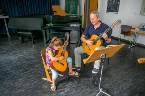 Mann und Mädchen, jeweils mit Gitarre sitzend, im Vordergrund ein Notenständer, im Hintergrund ein Klavier