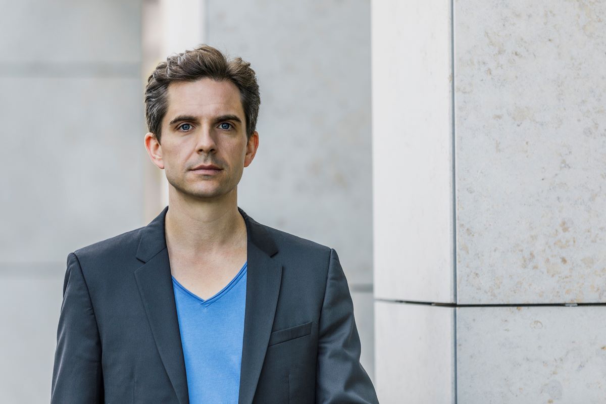 Junger Mann mit blauem Shirt unterm Sakko vor grauer Wand