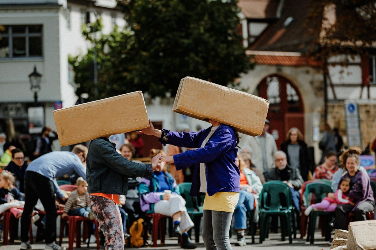 Theaterperformance im öffentlichen Raum, zwei Personen tragen je einen Karton über dem Kopf