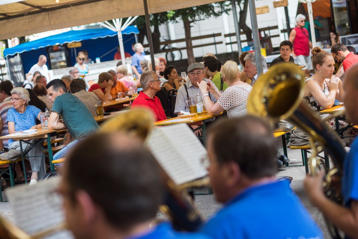 Menschen sitzen unter Schirmen auf Bierbänken. Im Vordergrund spielt eine Blaskapelle