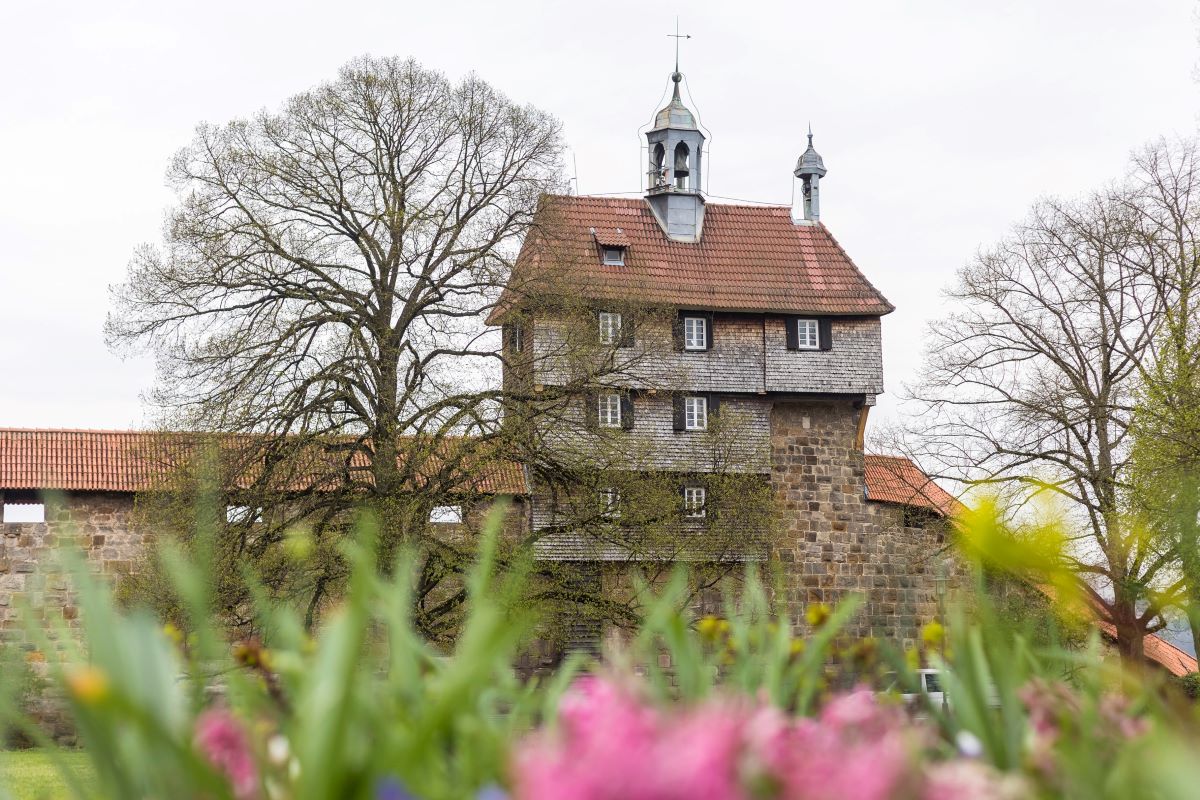 Schindelbedecktes Gebäude mit Glockentürmchen auf einer Mauer. Im Vordergrund Blumen.