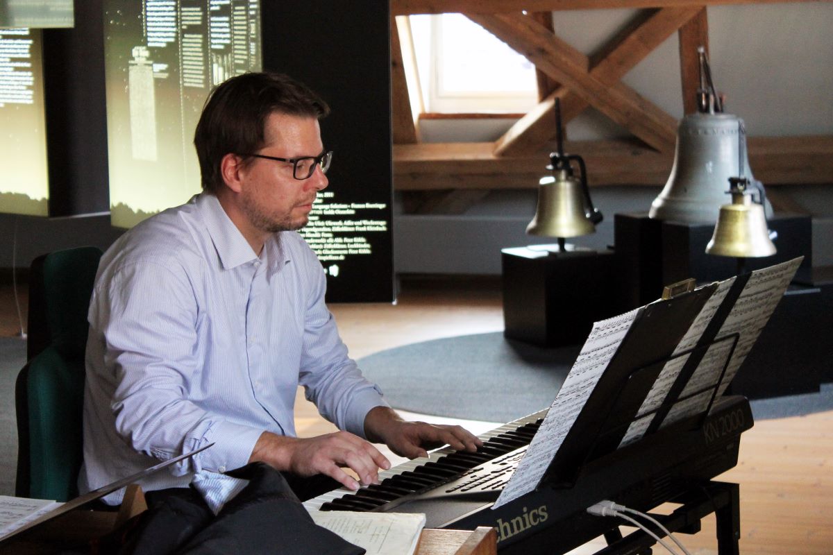 Mann mit Hemd und Brille an einem Keyboard. Im Hintergrund des Raumes sind Glocken in verschiedenen Größen zu sehen