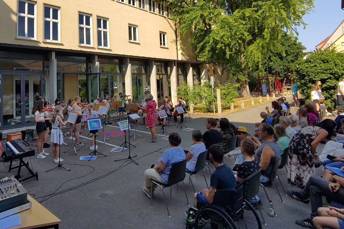 Vor einem Gebäude im Schatten spielt ein Kinder-Streichorchester, gegenüber sitzt Publikum auf Stühlen.