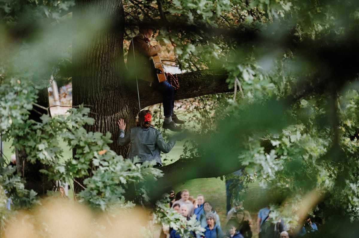 Künstler:innen sitzen in einem Baum, Publikum steht im Park darunter