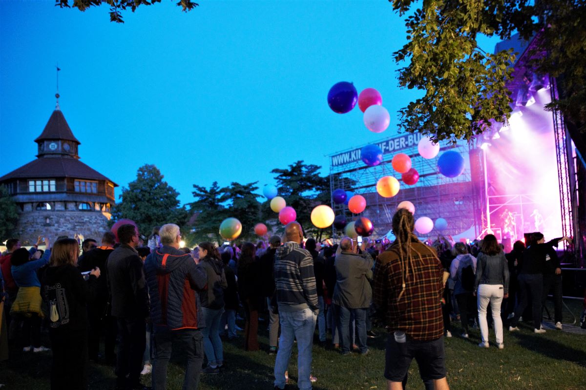 Menschen auf einer Wiese im Freien. Links der Dicke Turm der Esslinger Burg. Rechts eine Konzertbühne mit Scheinwerferlicht. Der Himmel ist nachtblau. Über dem Publikum schweben bunte Bälle.