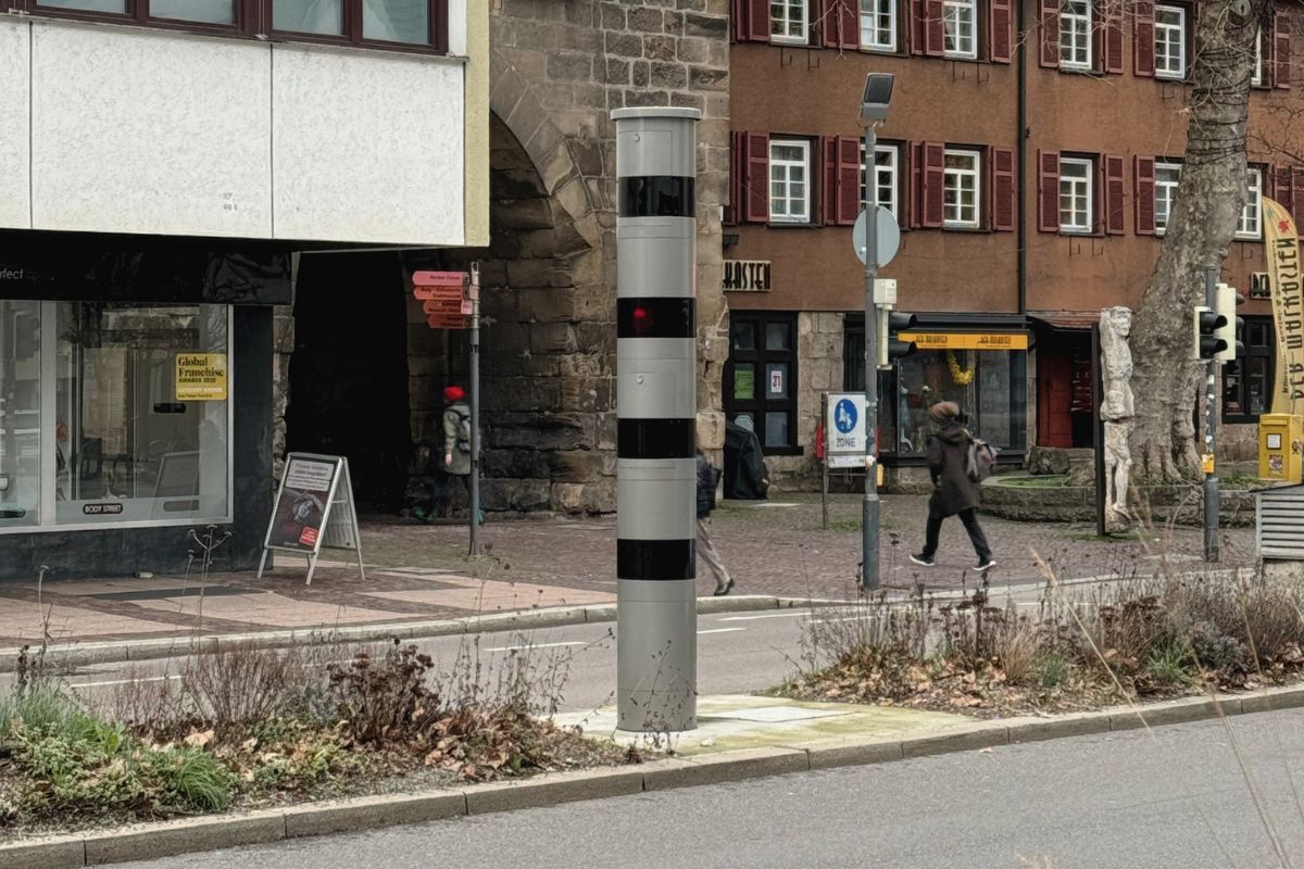 Grau-schwarz gestreifte Blitzersäule steht auf der Mittelinsel einer Straße