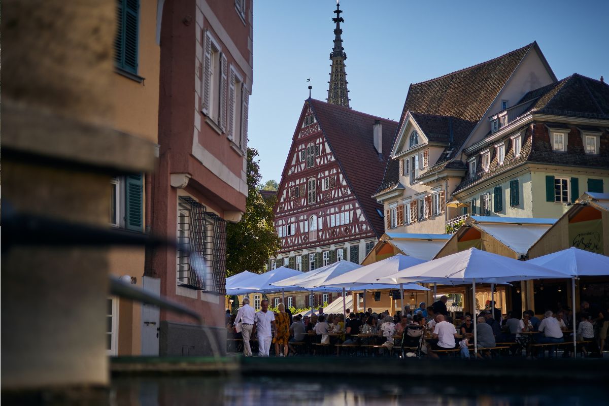 Vor der Fachwerkkulisse in Esslingen stehen Holzlauben und große Schirme. Auf den Bänken sitzen viele Menschen.