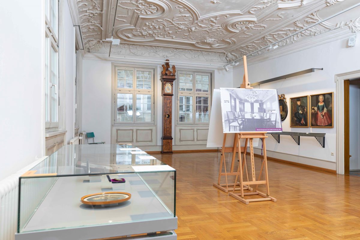 Blick in ein großes Zimmer mit Stuckdecke, an der hinteren Ward zwei große Sprossenfenster, in der Mitte des Raums steht eine Staffelei, links eine Vitrine