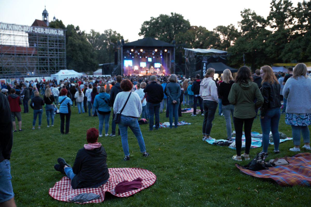 Menschen stehen oder sitzen auf Picknickdecken auf einer Wiese vor einer Konzertbühne. Im Hintergrund ist die Hochwacht der Esslinger Burg zu sehen