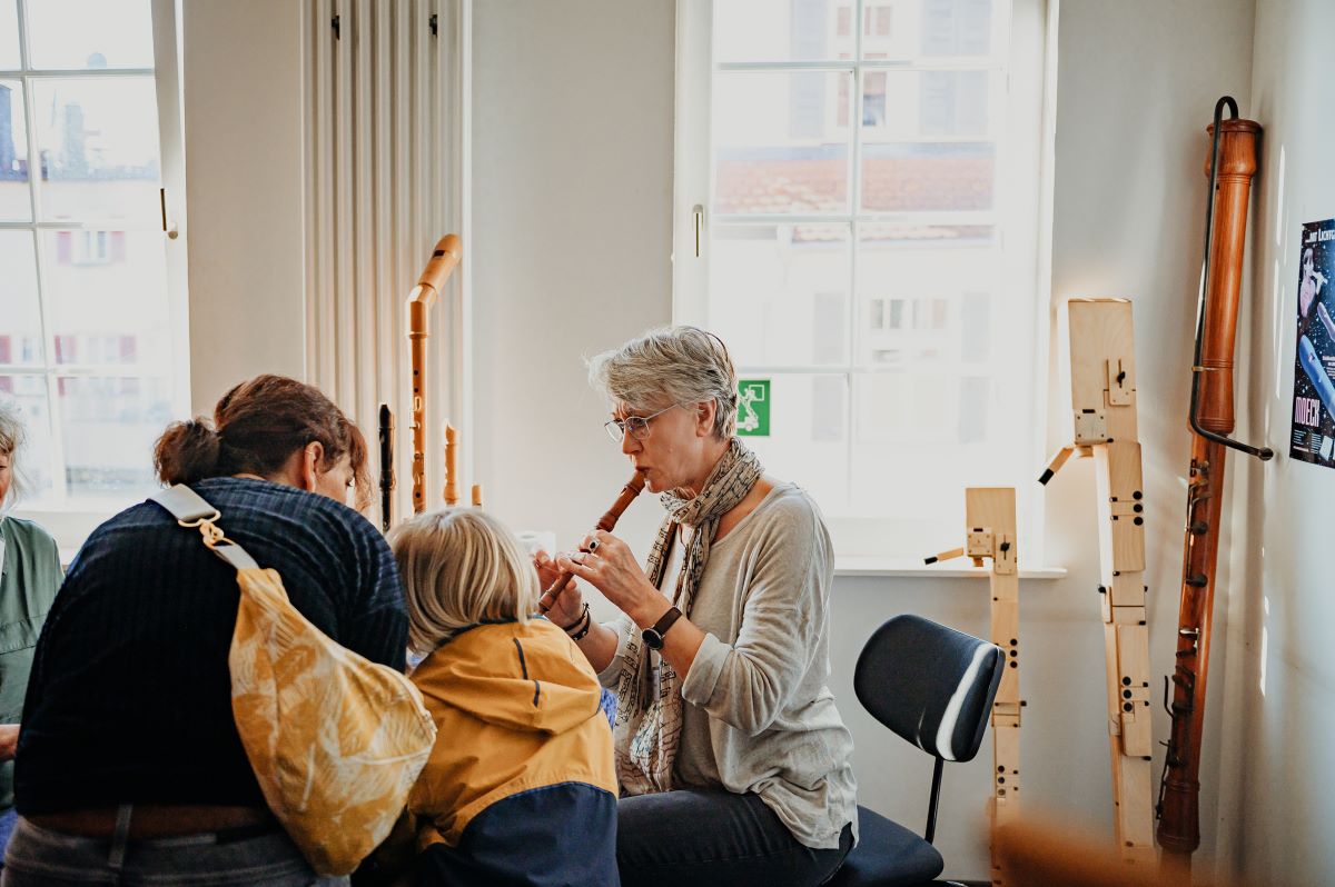 helles Zimmer mit vielen Holzflöten. Eine ältere Frau spielt Blockflöte. Eine Kind und eine jüngere Frau sehen und hören zu