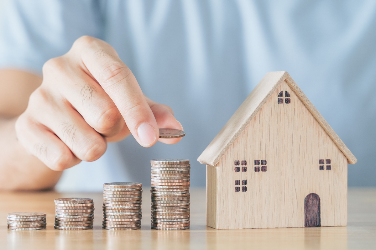 Haus mit Geld Eine Person stapelt Münzstapel neben einem kleinen Holzhaus